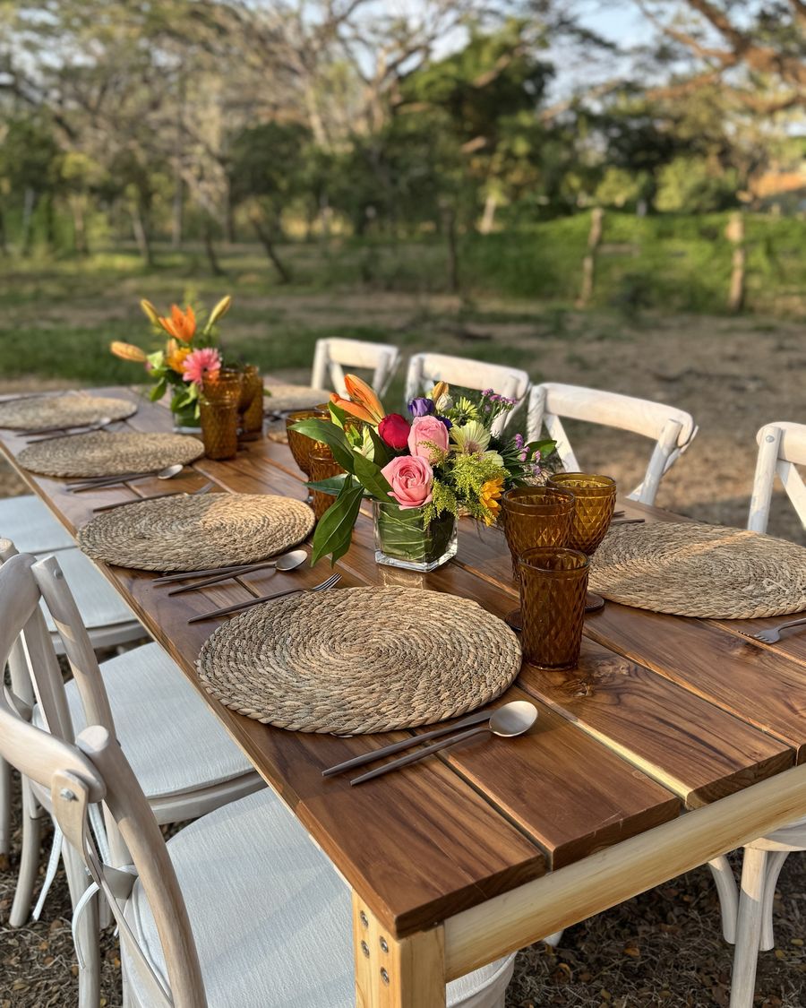Teak table set with woven wicker chargers, amber glassware and fresh flowers, with white crossback chairs, outdoors in Costa Rica